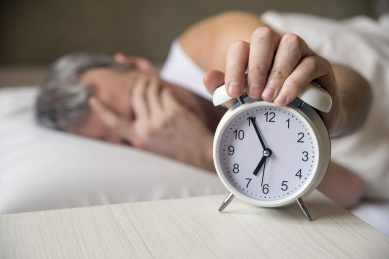 Hombre tumbado en la cama tapandose la cara y apagando el despertador